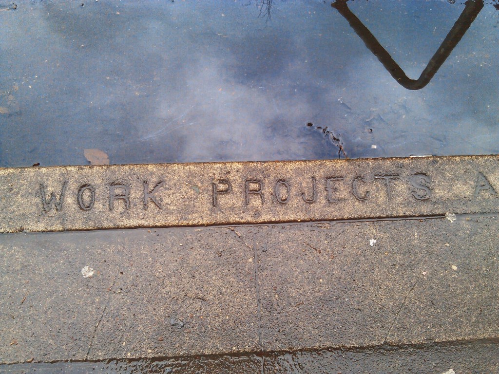 A wet sidewalk and street closeup with letters in the cement curb that read "Work Projects," and an "A," so part of "Works Projects Administration," from the Great Depression years. 